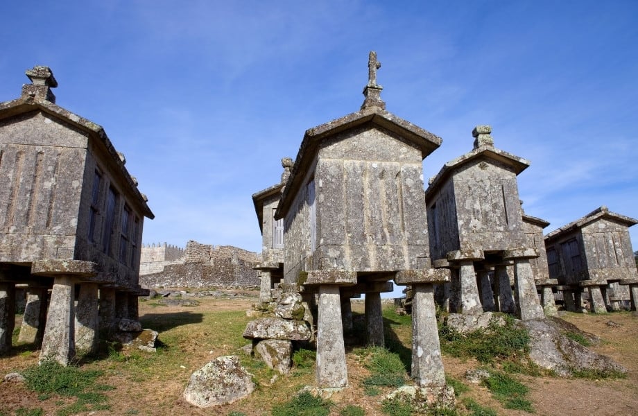 Places to visit in northern Portugal, stone single-storey buildings on stone stilts sat atop grassy area in front of old fortification ruins under a blue sky with wispy white clouds