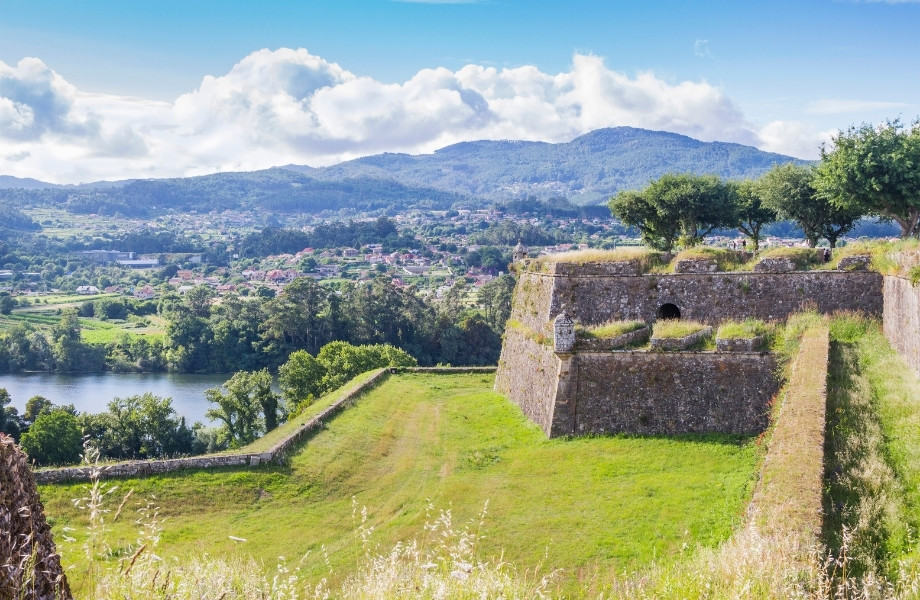 What to do in north Portugal, moss-covered stone walls with grass areas and green trees with river running alongside and forested village with mountain in the distance under a cloudy sky