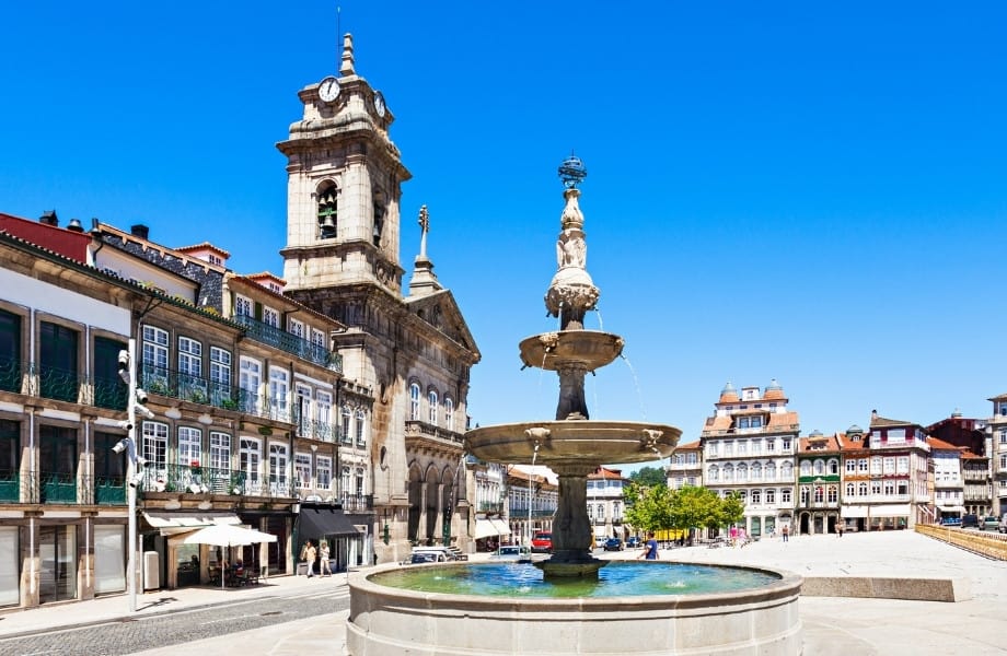 best places to visit in northern portugal, ornate three tiered fountain in open square surrounded by three-storey buildings with balconies and a large stone bell tower with four clocks under an azure blue sky