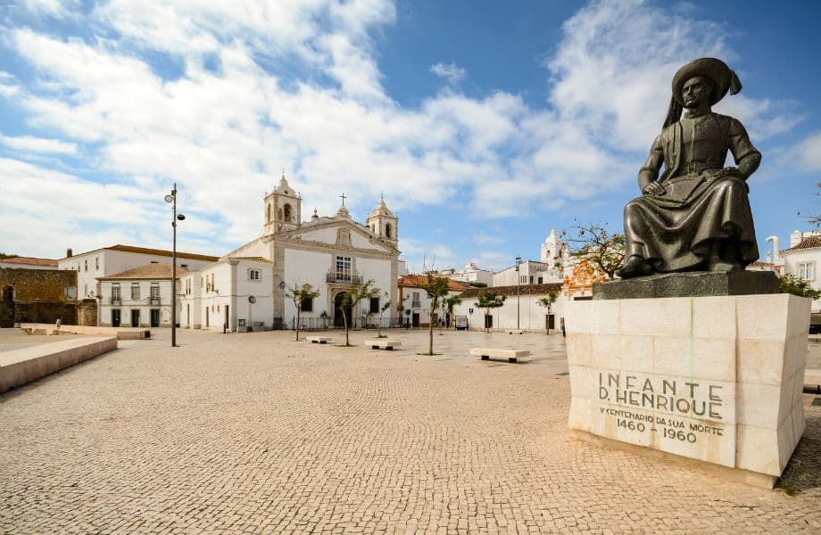 must see places to visit in Lagos Portugal, large courtyard in front of a church with a stone statue at forefront of a sitting man with a wide hat