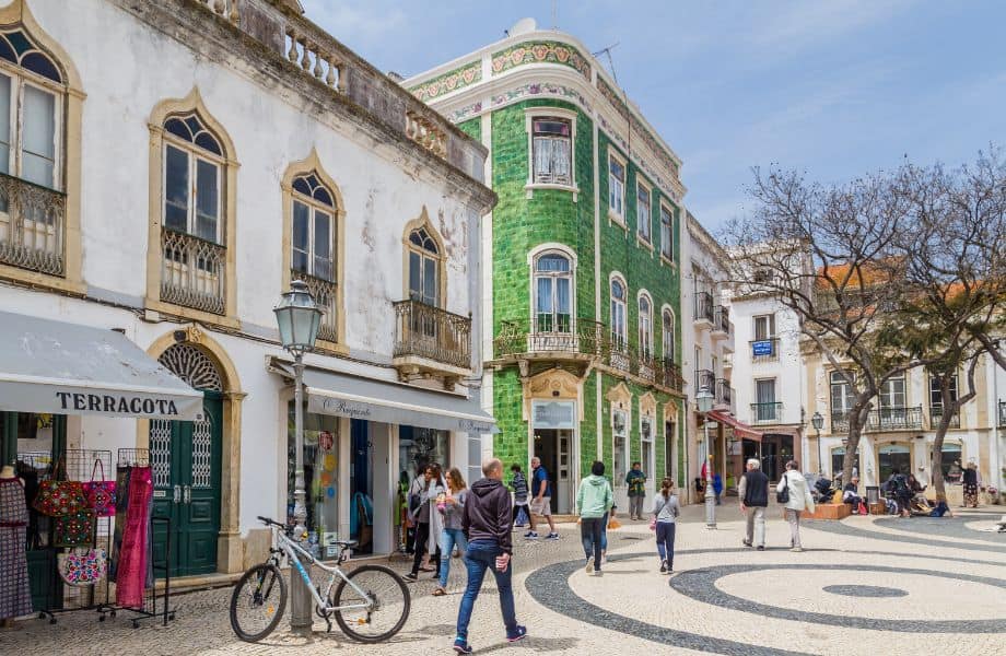 people walking through large open stone square next to old white building with awnings and tall green tiled historic building