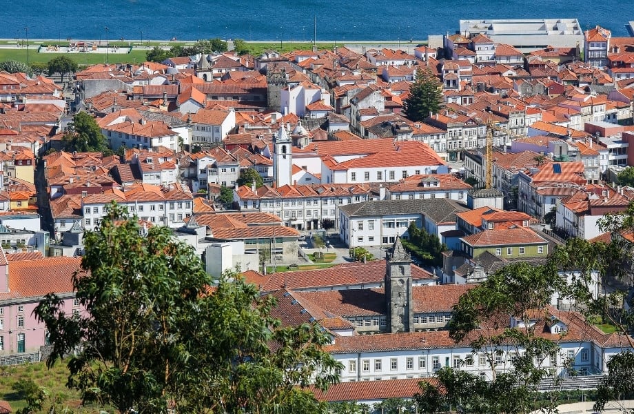 What to see in northern Portugal, view overlooking the terracotta rooftops of many old buildings laid out in irregular patterns with streets winding in-between with blue sea in background and green trees in foreground