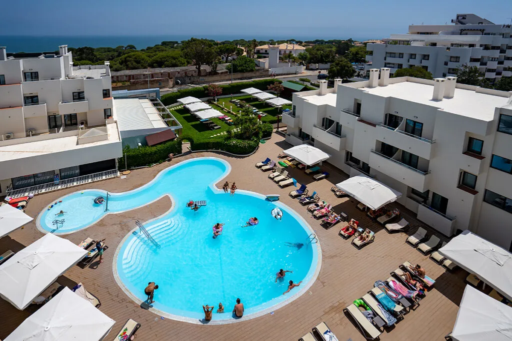 Albufeira family hotels, aerial view of oddly shaped hotel pool with people swimming and surrounded by people suntanning in pool chairs and white buildings with beach in distance