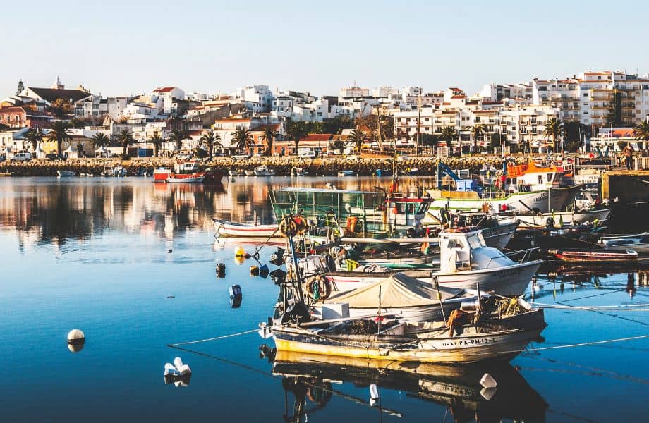 view of town with many white buildings from water, many small fishing boats in water between photographer and town, Where To Stay In Algarve For Nightlife