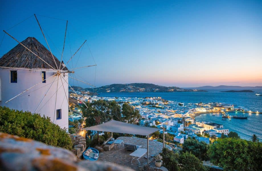 view of Mykonos coastal city lit up at sunset with private covered patio and windmill in foreground and mountains in the distance