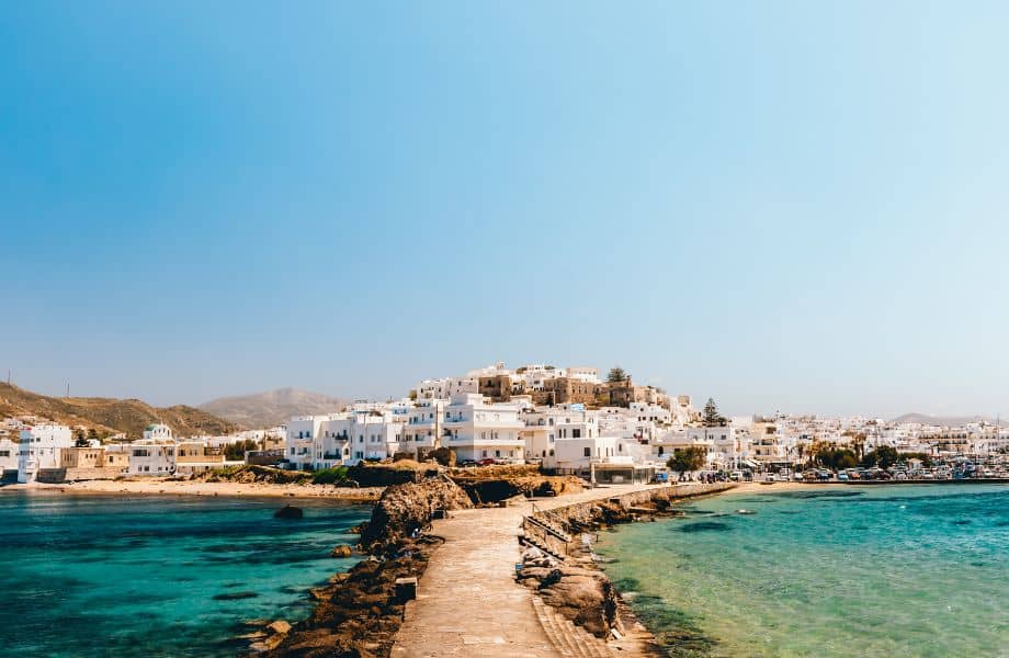 how to get to Paros Island from Naxos, coastal hillside city covered with mostly white and blue buildings on a clear day surrounded by clear blue water and seaside path heading into the water with mountains in the distance