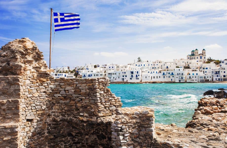 Exactly How to Get to Paros, Greece, image white and blue buildings lined up along the coast with clear blue water and in foreground stone ruins with a Greek flag sticking out