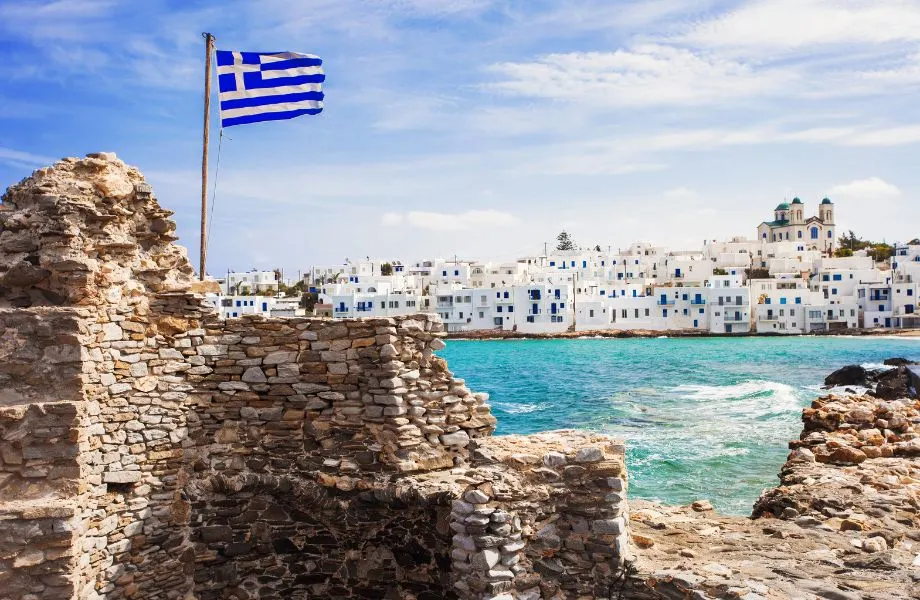 how to get to Paros Exactly How to Get to Paros, Greece, image white and blue buildings lined up along the coast with clear blue water and in foreground stone ruins with a Greek flag sticking out