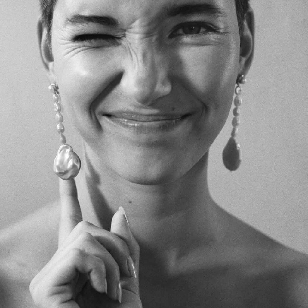 luxury ethical jewelry brands, black and white image of person showing off dangly earrings and making a playful face