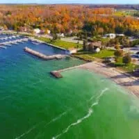 aerial view of a coastal bay town surrounded on one side with bright green and blue waters and and on the other with miles of orange and yellow trees on a sunny day, find here the best hotels in Sister Bay