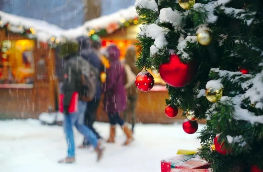Top Christmas markets in Wisconsin, people walking in snow in front of market stalls in background with corner of Christmas tree in forefront