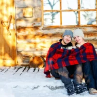 couple sitting outside a log cabin in the snow underneath a blue and red checkered blanket at one of the best romantic cabins in Wisconsin Dells