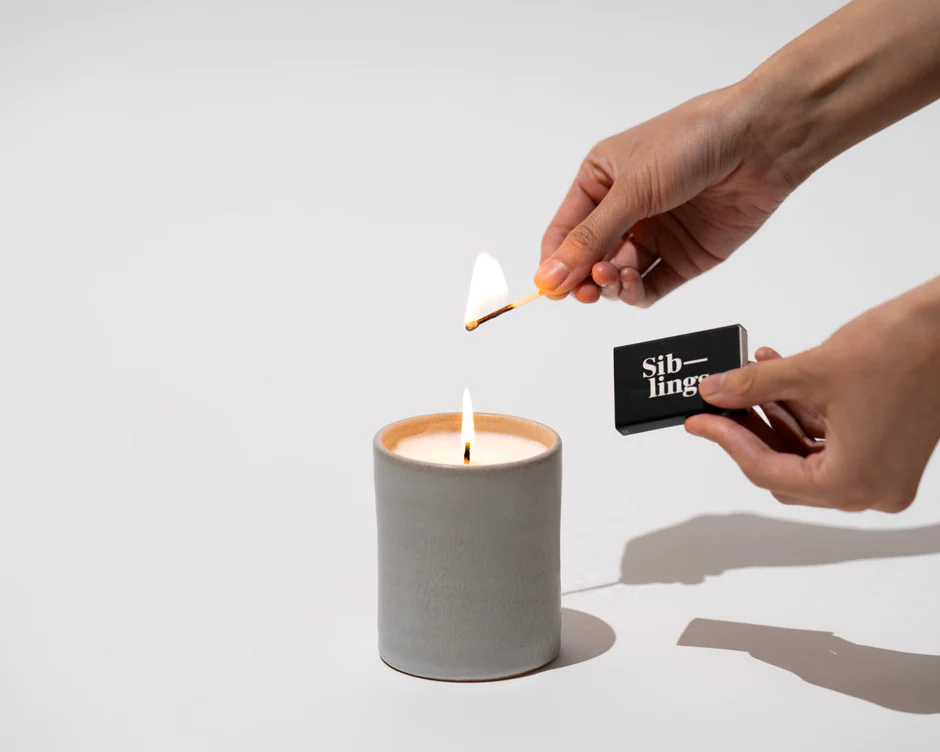 cool eco-friendly gifts, person lighting candle in ceramic holder with matchbook reading 'siblings'