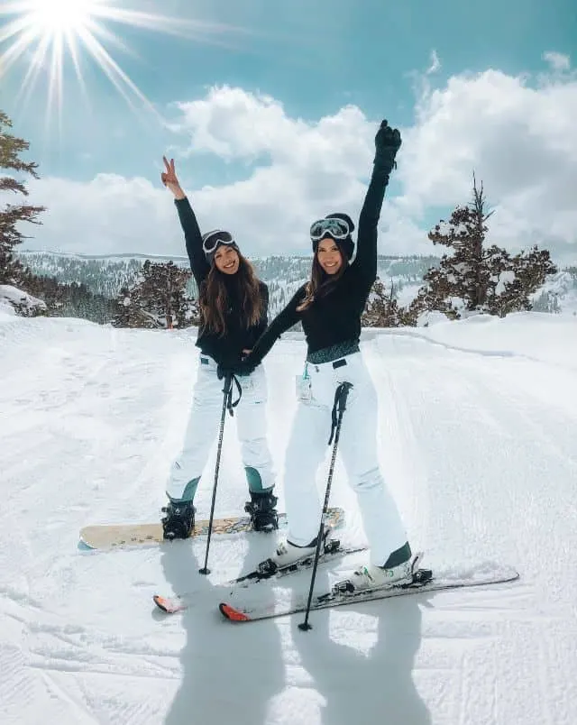 two women standing on a ski slope on a bright sunny winter day with a hand extended in the air and smiling, one woman is in skis and the other is on a snowboard