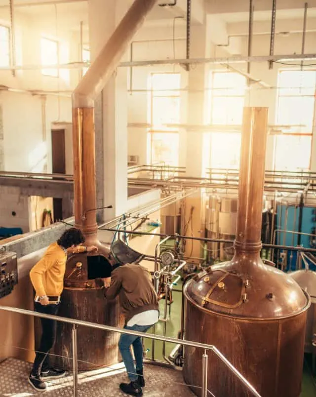 Plan a trip to the best craft breweries in Milwaukee, two people examining a beer brewing machine in a brewing factory with sunlight pouring in through large windows behind