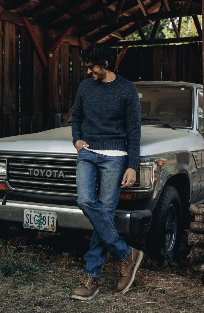 best sustainable denim brands for men, white man wearing blue sweater and blue jeans leaning against a car