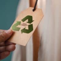 Best zero waste fashion brands, person reading tag on shirt that reads 'made by 100% recycling materials' inside of a shiny green recycle symbol