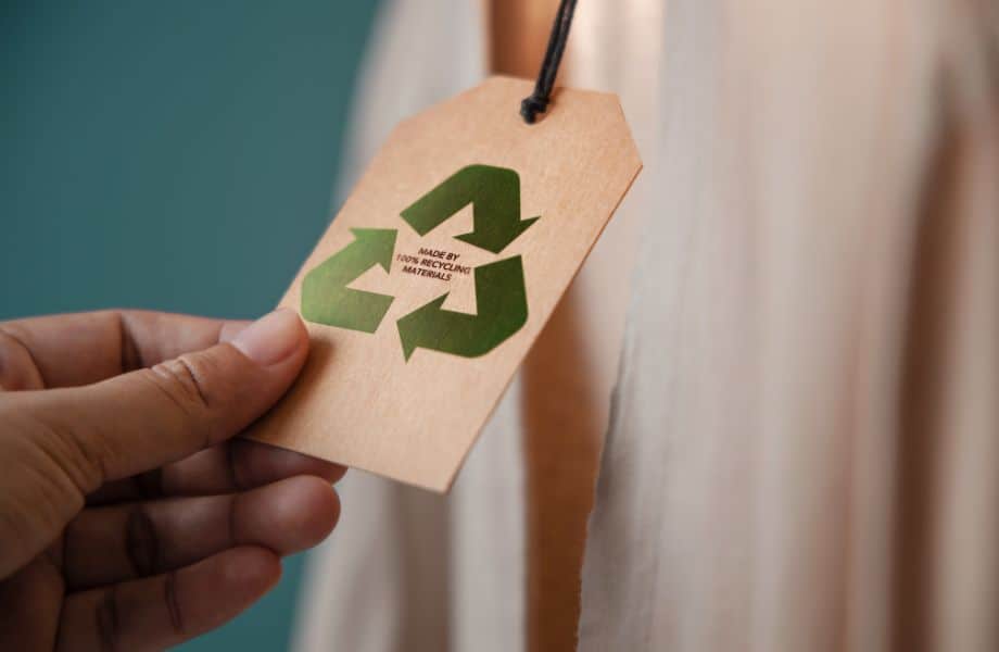 Best zero waste fashion brands, person reading tag on shirt that reads 'made by 100% recycling materials' inside of a shiny green recycle symbol