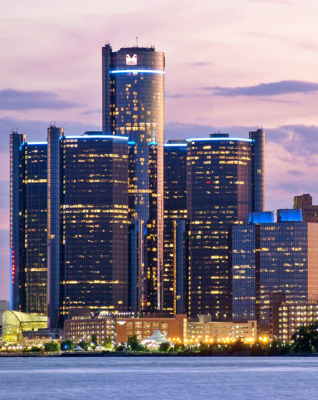best road trips from Milwaukee Wisconsin, buildings in downtown Detroit with tark tinted windows lit up at dusk under a purple sky next to calm water