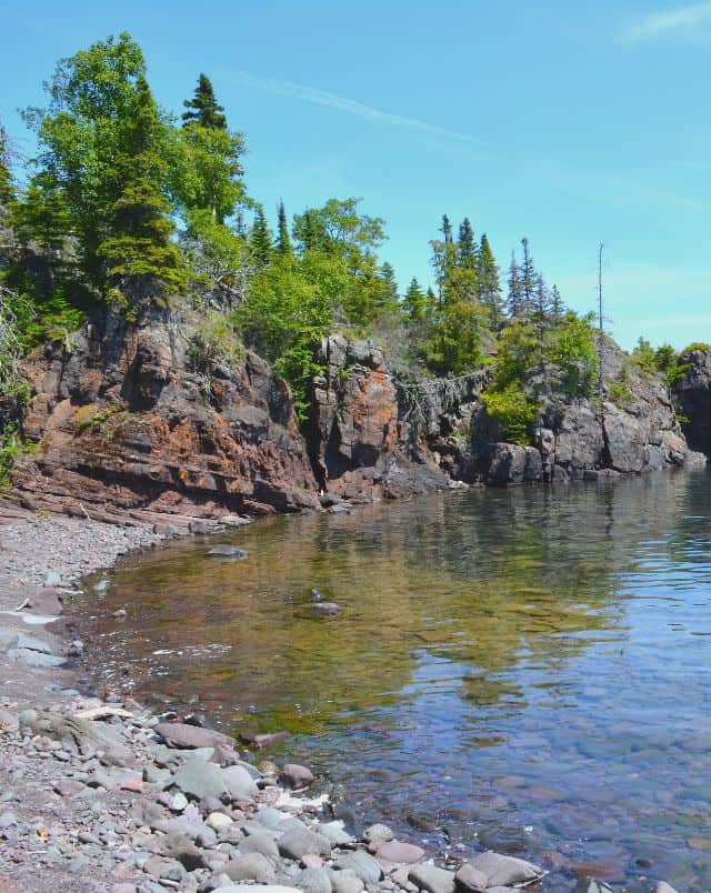 best road trips from Wisconsin, rocky coast next to clear water with taller jagged rocks covered by trees further along the shore