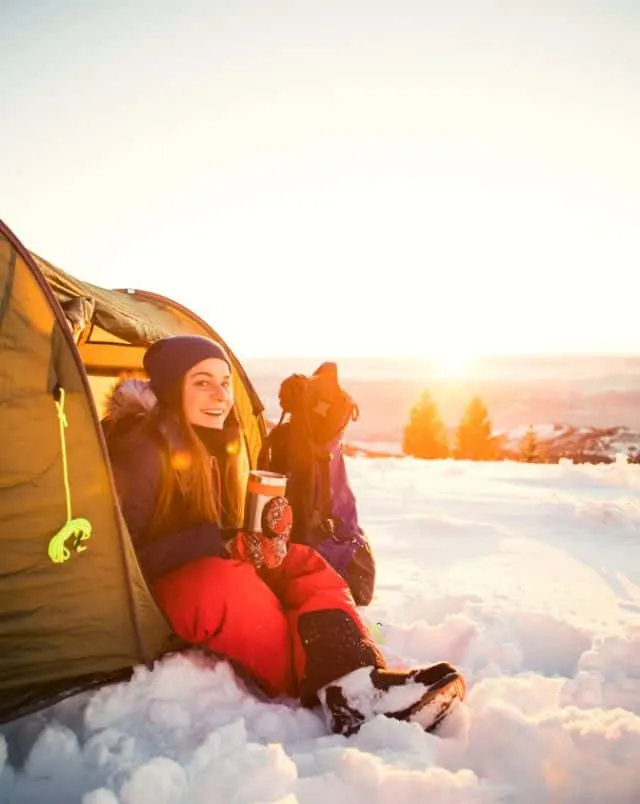 best places for winter camping in Wisconsin, person sitting half in a tent surrounded by snow, half in snow, holding a metal mug with rising sun in background