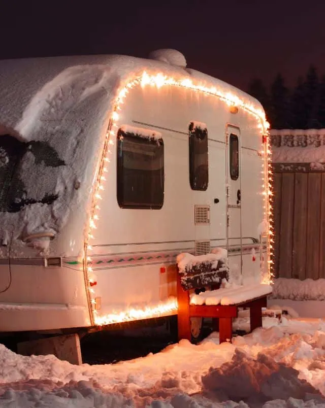 best winter camping in Wisconsin, lit up RV covered in snow parked next to wooden bench also covered in snow
