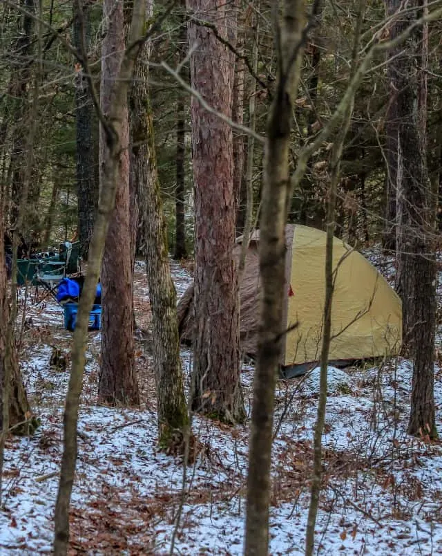 where to go camping in the winter in Wisconsin, tent and camp gear set up on ground with light dusting of snow behind many trees