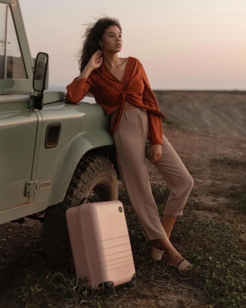 best eco-friendly suitcases, biracial woman leaning against green jeep with pink hardshell suitcase next to her and dusk