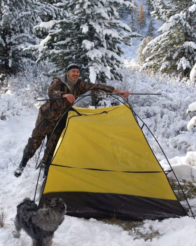 snowy camping in Wisconsin, person putting up a tent in the snow while a dog watches with snow covered trees behind