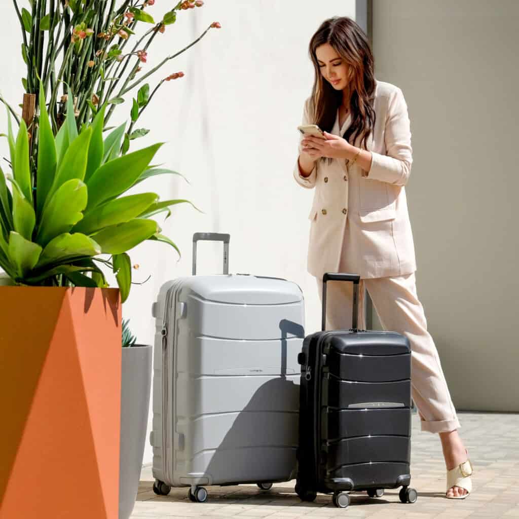 non toxic luggage brands, Asian woman in tan suit standing next to two suitcases while looking at her phone