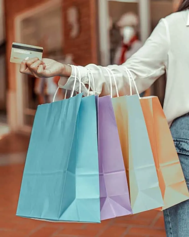 person holding several paper shopping bags of different colors on outstretched arm with a credit card in their hand