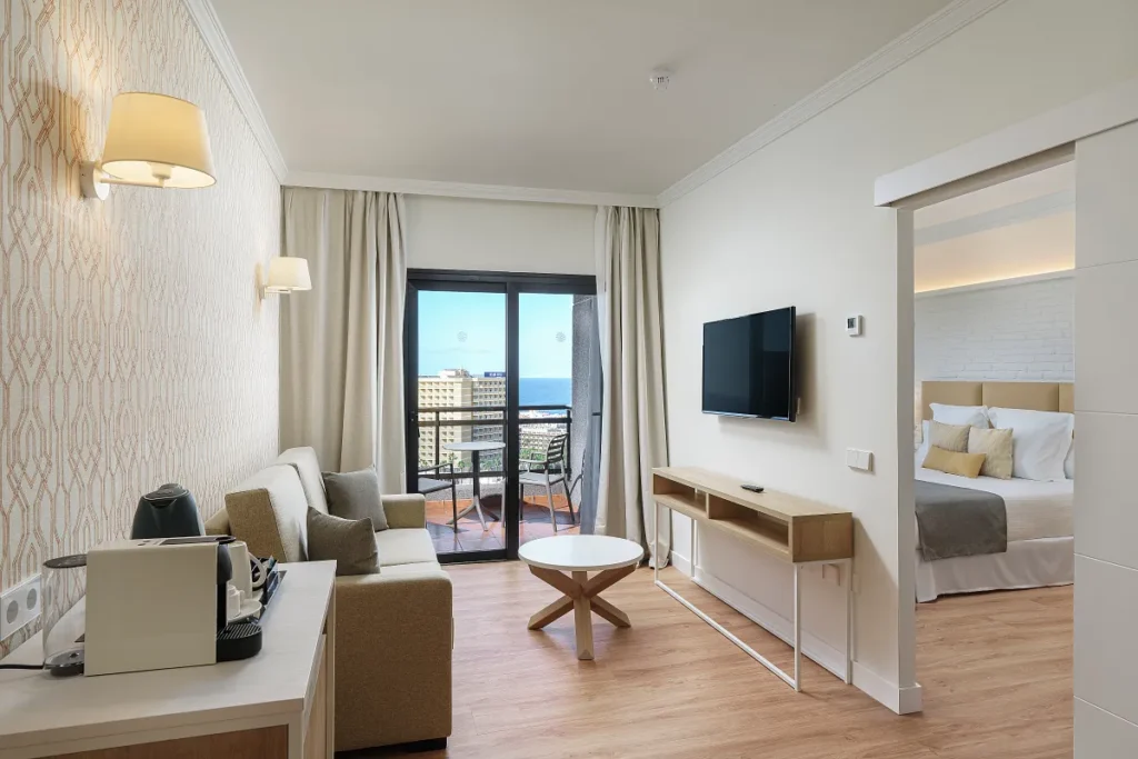 best hotels in Puerto de la Cruz hotels for luxury stays, Modern hotel room with cream coloured walls and colour coordinated tables and sofa with balcony visible through glass door and bedroom off to one side