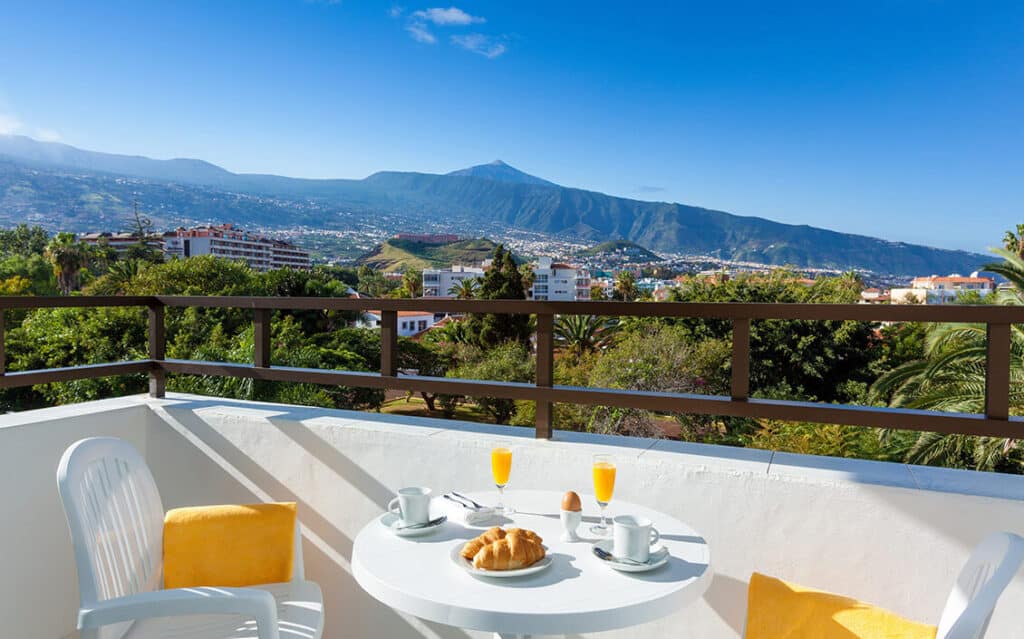best hotels Puerto de la Cruz Tenerife has to offer, white balcony with white plastic chairs and a white table set with mugs, flutes of orange juice, plate of croissants and a poached egg, the view from the balcony is a lot of greenery, behind which peeks out a sprawling city with mountains in the distance under a clear blue sky