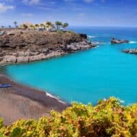 Resorts in South Tenerife, View of sandy beach next to vibrant sky blue water with rocky coastline to one side upon which sits a complex of buildings with palm trees outside all under a deep blue sky with white wispy clouds