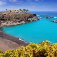 Resorts in South Tenerife, View of sandy beach next to vibrant sky blue water with rocky coastline to one side upon which sits a complex of buildings with palm trees outside all under a deep blue sky with white wispy clouds