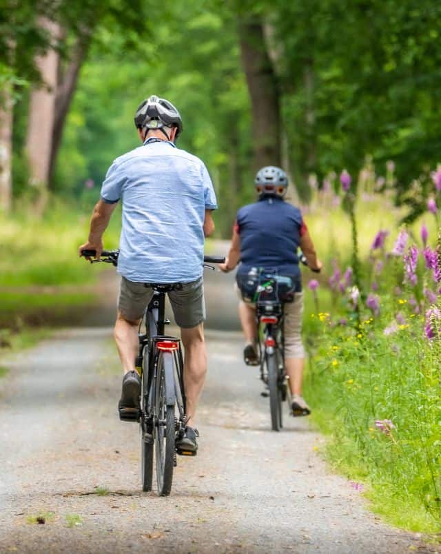 best activities in Milwaukee to do outdoors, older couple cycling away from camera on a work bike bath surrounded by trees, grass and purple flowers