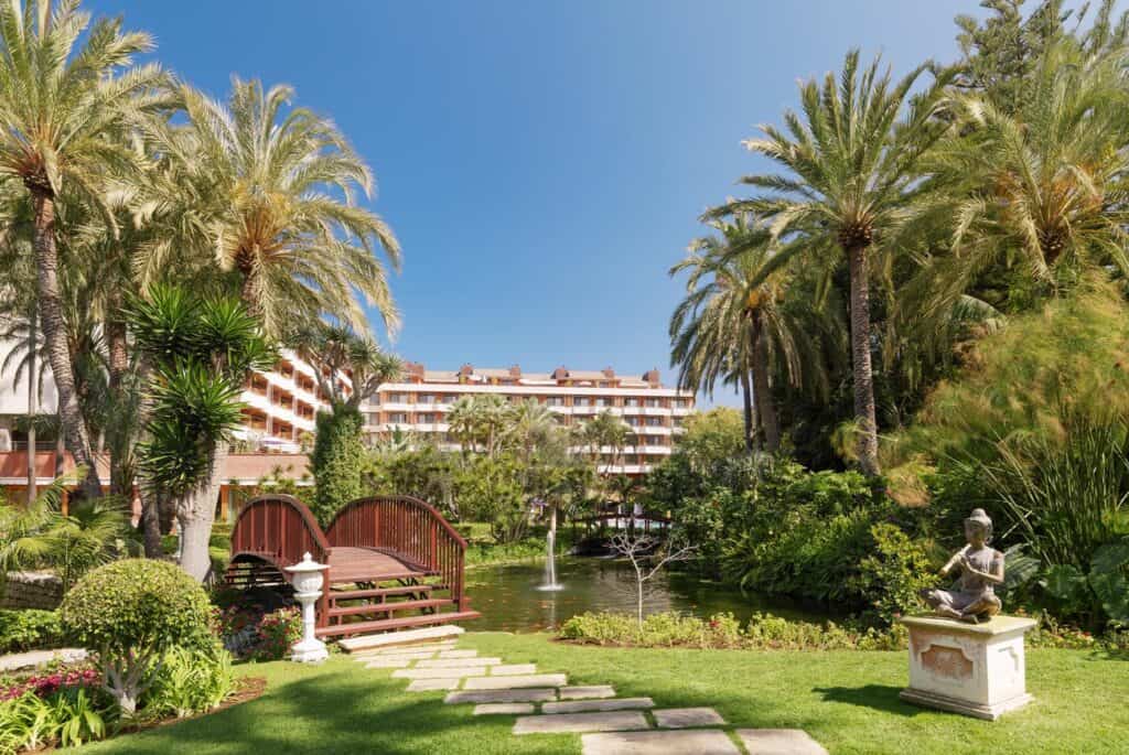 5 star hotels in Puerto de la Cruz, Hotel garden area with neatly trimmed grass lawn surrounding paved stone steps leading to a small wooden bridge crossing over an idyllic stream and pond with a small fountain all surrounded by tall palm trees underneath a bright blue sky