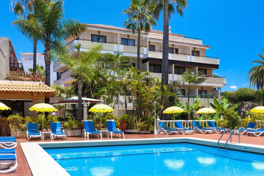 best Tenerife Puerto de la Cruz hotels, hotel pool with hotel and trees behind under a blue sky, pool is lined with blue pool chairs and yellow umbrellas