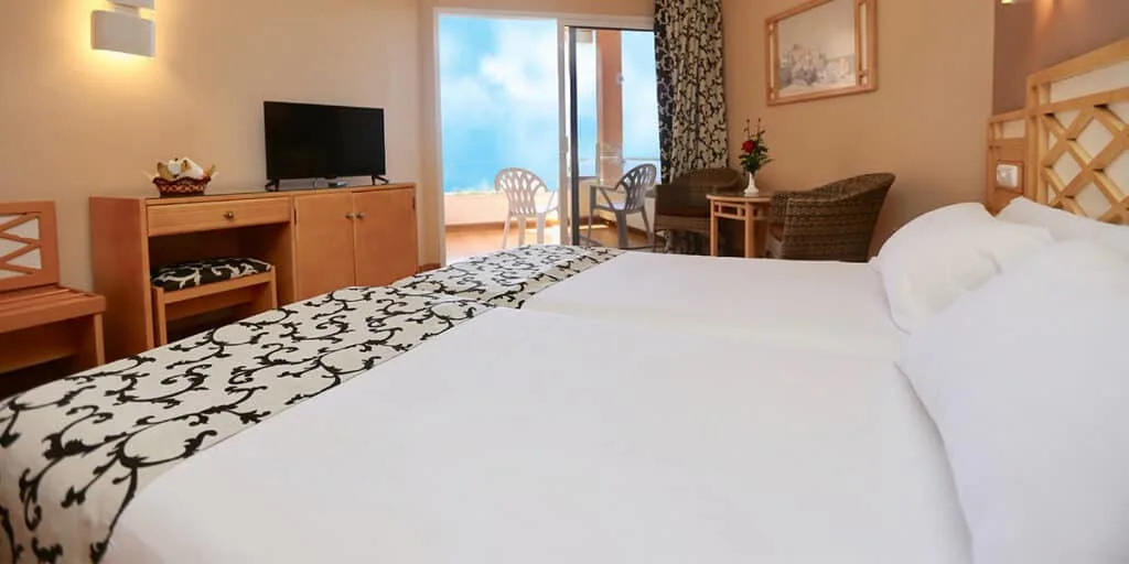 Puerto de la Cruz Tenerife hotels, inside of hotel room with two beds pushed together to make one, in front of bed is a table with a TV and to the side wicket chairs and a table, beyond this is a balcony with white plastic chairs and a view of a bright blue sky
