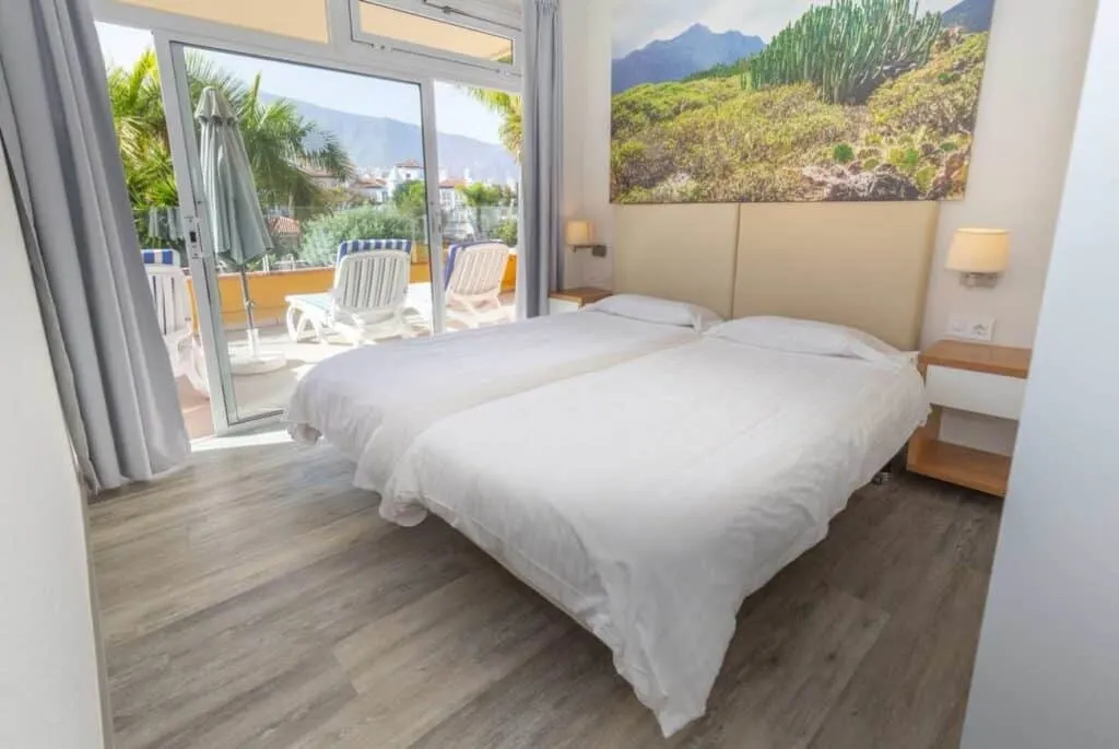 where to stay in Tenerife in winter, Hotel bedroom with large twin beds covered in pristine white bedsheets and large picture of mountain foliage overhead with a glass door leading to an outdoor balcony area complete with several sun lounger chairs and a view of green trees and buildings in the distance