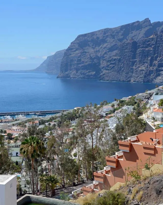 View from hillside populated with buildings and small trees out across the sea with deep blue water next to large rocky coastline cliffs under a hazy clear sky, Where To Stay In Tenerife For Hiking at Los Gigantes 