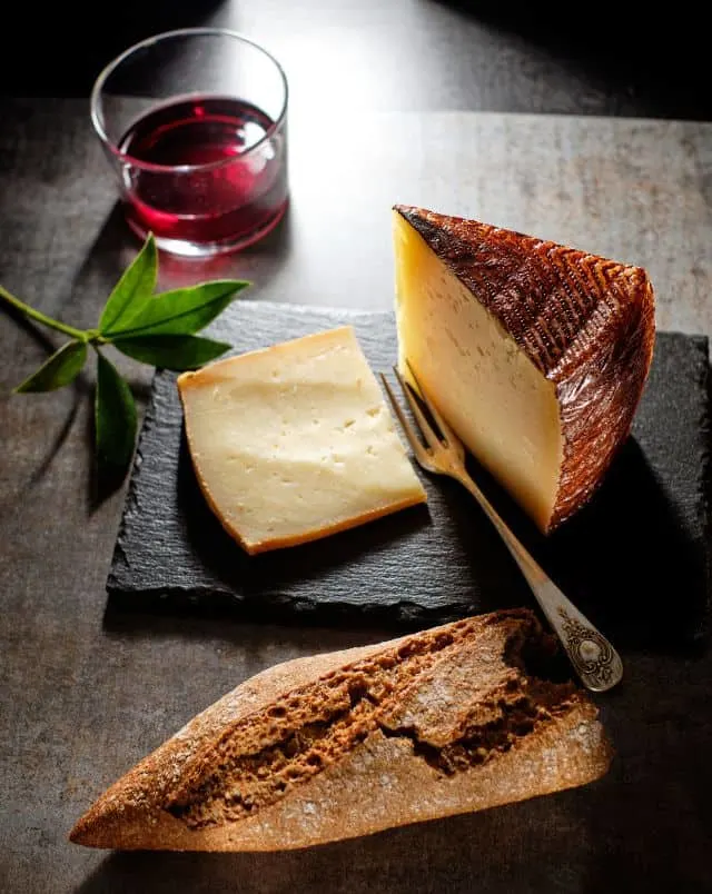 large chunk of Manchego Cheese with a slice taken out sitting on a black slate next to a metal fork and a half loaf of crusty bread