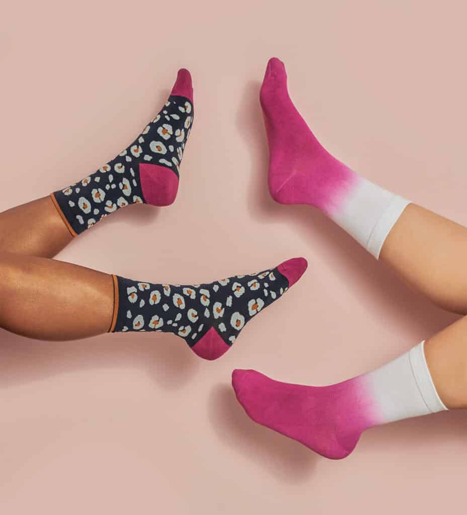 ethical bamboo socks, socked feet of two people holding up their feet against a pastel pink background, socks of person on right are pink on the feet and fade to white on the ankle, socks of person on left are black with white flowers and pink heels and toes