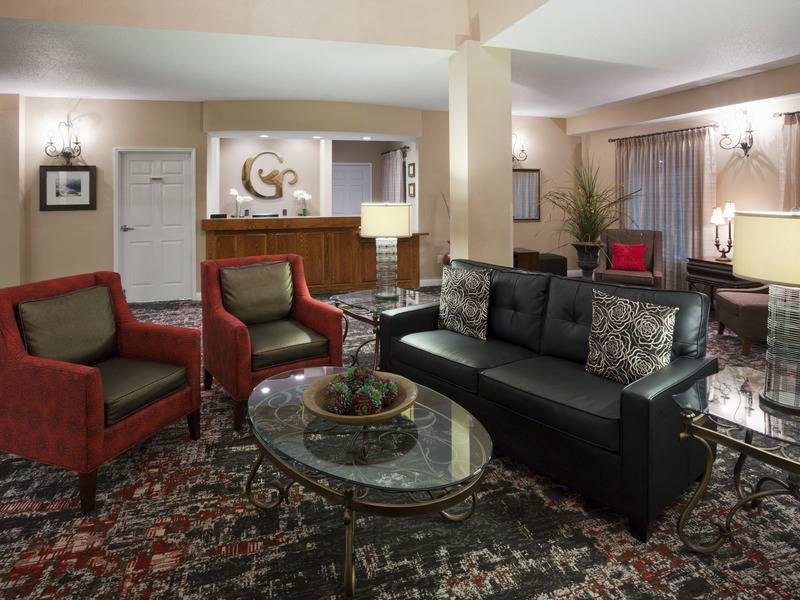 Hotel lobby with low ceiling populated by a mixture of styles of furniture including a black leather sofa and red stiff armchairs surrounding a glass table with other grey armchairs behind and a reception desk area behind