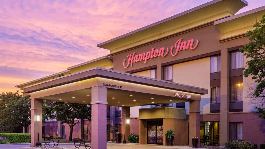 Large front porch area of hotel with covered entryway and park benches leading to sliding electric doors all under large illuminated lettering that says "Hampton Inn" in pink-tinted cursive script all under a cloudy sky at sunset