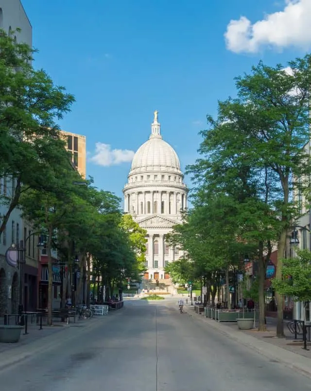 family weekend getaways in Wisconsin, view of Capitol building at the end of a street of shops and cafes that is empty except for a person a bike under a bright blue sky with a few clouds