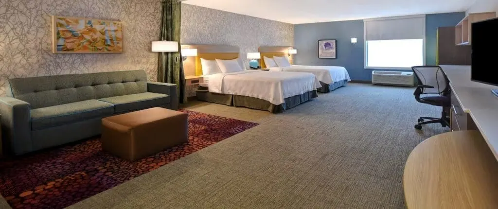 Very large long hotel room with wide sofa and footrest next to two wide double beds set next to each other opposite a long desk area with office chair and a tall window in the far wall