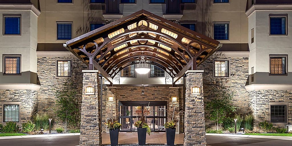 Front entryway to large hotel building with bespoke stone pillars holding up the wooden roof of a covered walkway with neatly arranged potted plants leading towards the inviting glass doors of the main entrance at nighttime