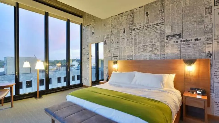 High-ceilinged hotel room with large double bed and floor-to-ceiling windows with a wall decorated in newspaper-style wallpaper with doorway leading into another area to one side