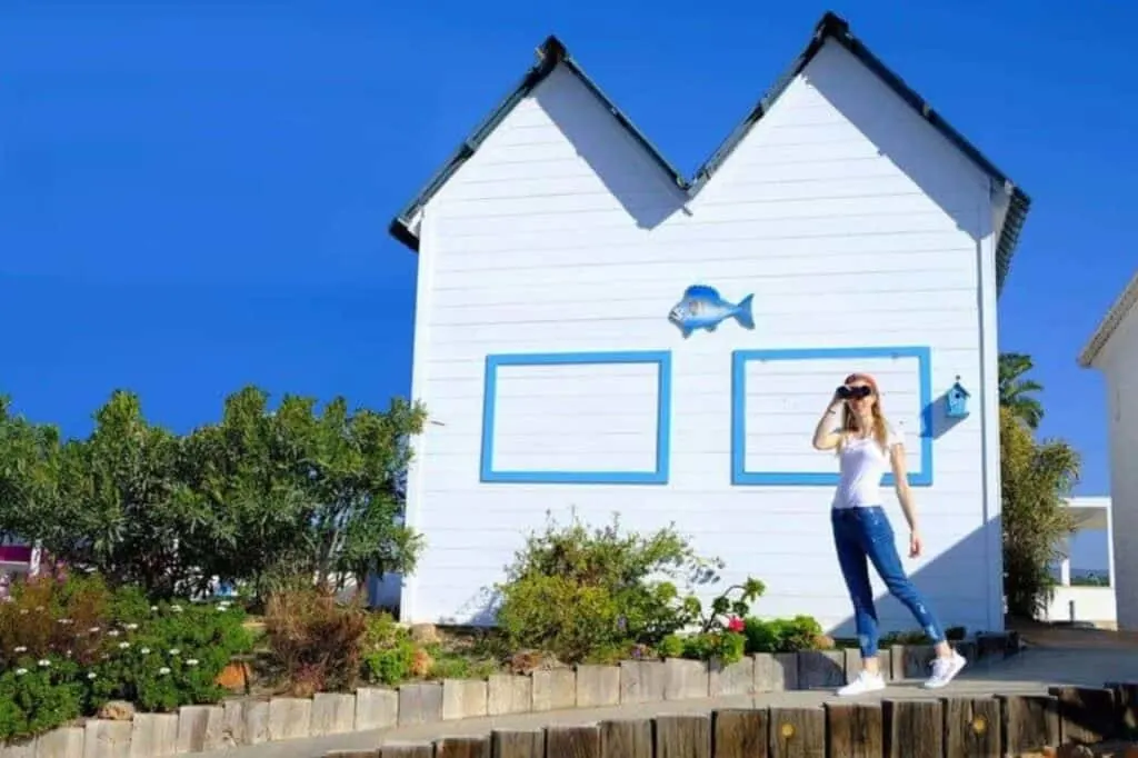 Blogger with binoculars in front of white and blue cottage near beach in Albufeira